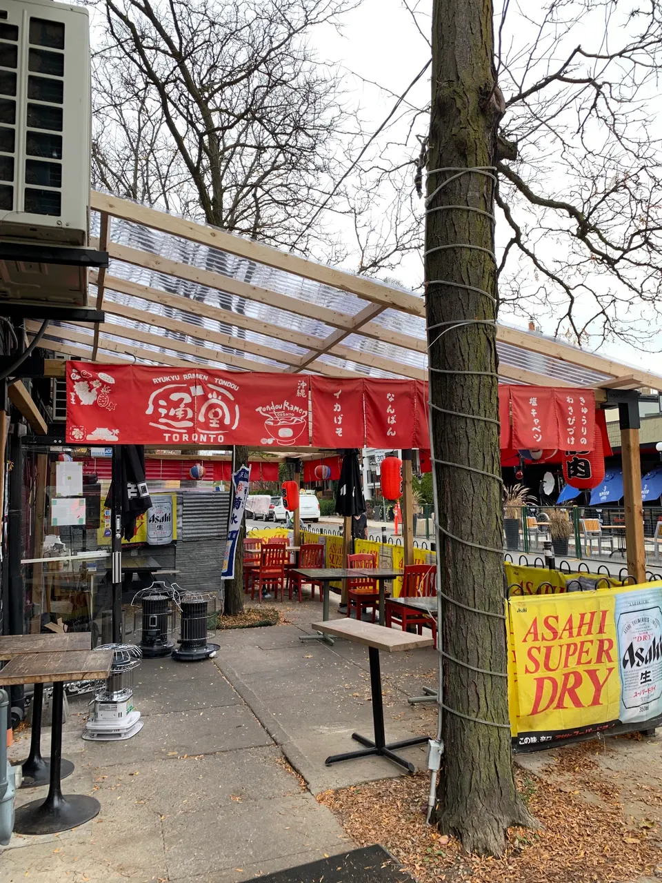 Outdoor patio of Tondou Ramen in Toronto, featuring a wooden pergola with a clear corrugated roof, red Japanese banners, outdoor tables and chairs, and an Asahi Super Dry banner.