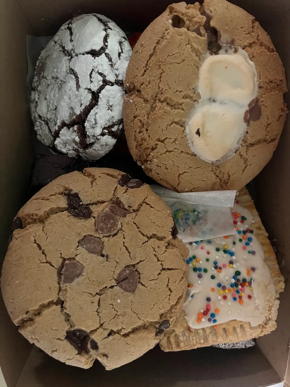 A cardboard box filled with four large baked goods, including a powdered chocolate crinkle cookie, a marshmallow-topped cookie, a chocolate chip cookie, and a frosted pop-tart style pastry with colorful sprinkles.