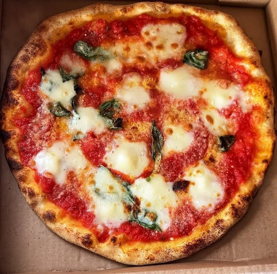 A freshly baked Margherita pizza with a charred crust, vibrant red tomato sauce, melted pools of fresh mozzarella cheese, and scattered green basil leaves, served inside a cardboard pizza box.