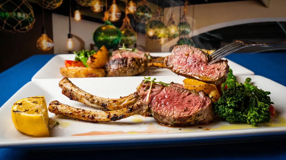 A close-up of grilled lamb chops served on a white plate with roasted potatoes, broccolini, and a grilled lemon wedge, with a fork lifting a tender, medium-rare slice of lamb.