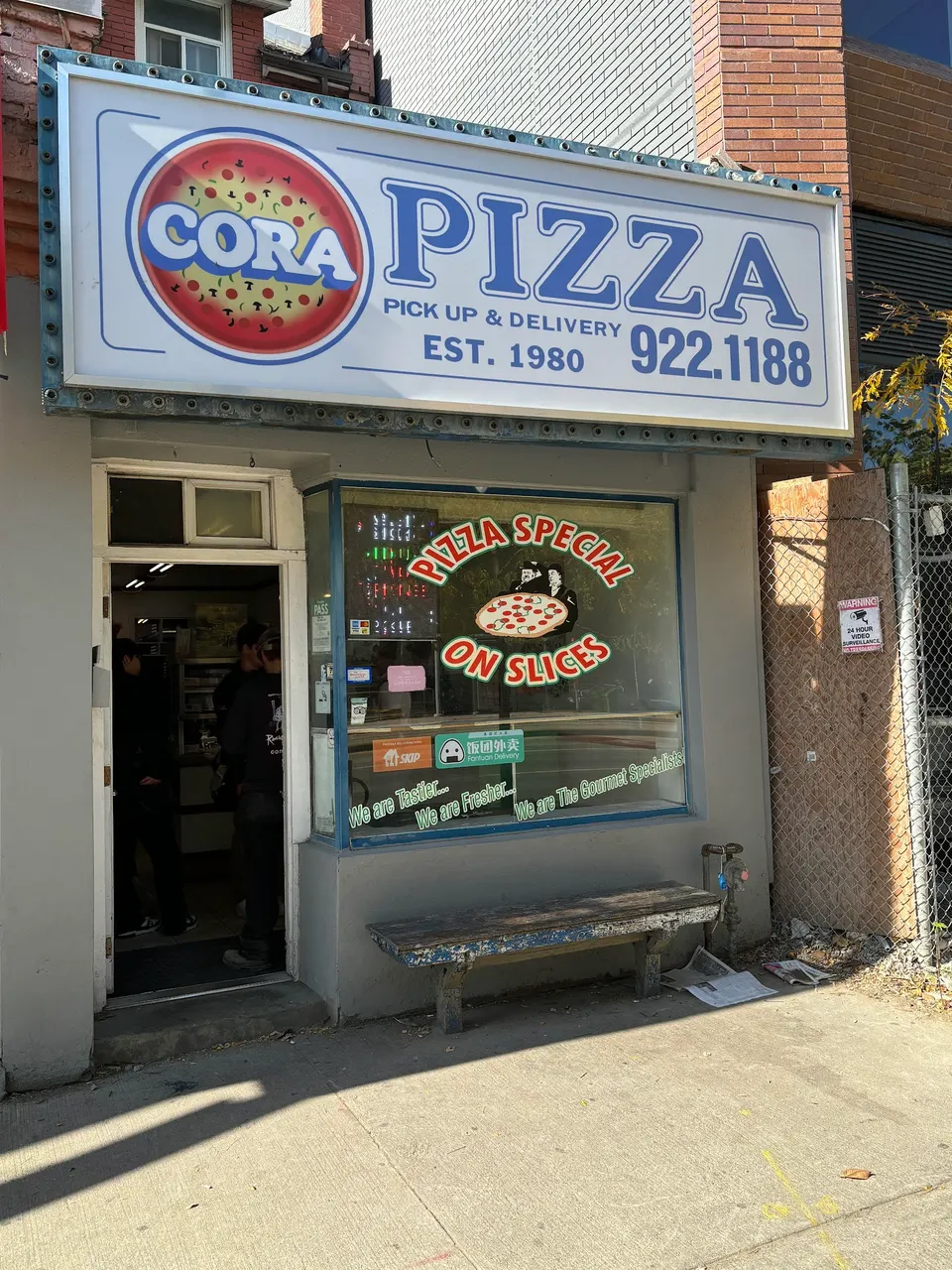 The storefront of Cora Pizza, featuring a large white sign with blue lettering, a glass window with 'Pizza Special On Slices' text, and a wooden bench outside.
