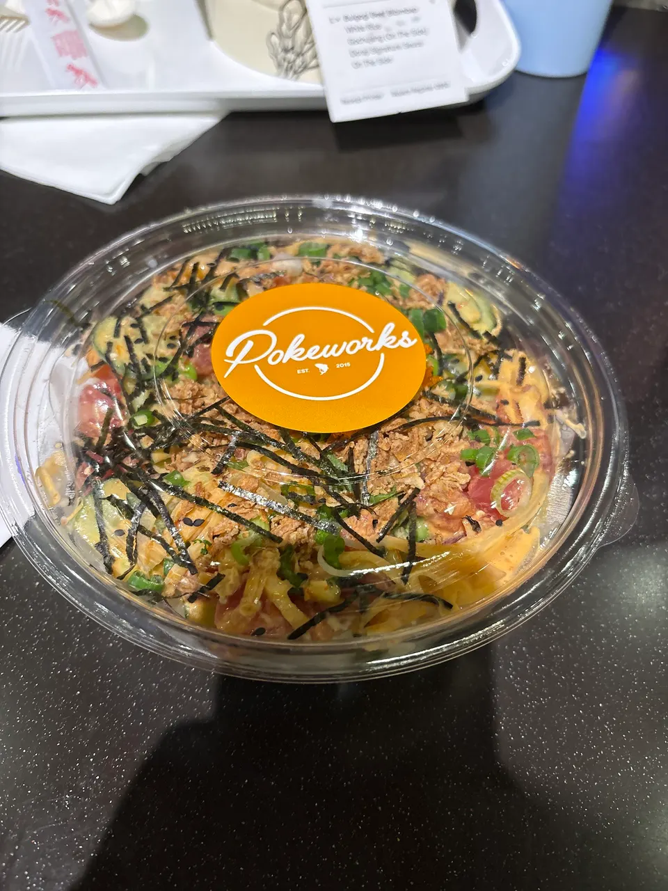 A clear plastic container filled with a poke bowl, topped with diced fish, sliced avocado, green onions, crispy fried onions, and seaweed strips, with an orange 'Pokeworks' logo sticker on the lid, sitting on a dark table.