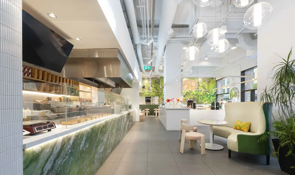 Modern interior of Green Box restaurant featuring a long green marble counter, white walls, high ceilings with exposed pipes, spherical pendant lights, and a cozy seating area with a curved green and beige sofa.