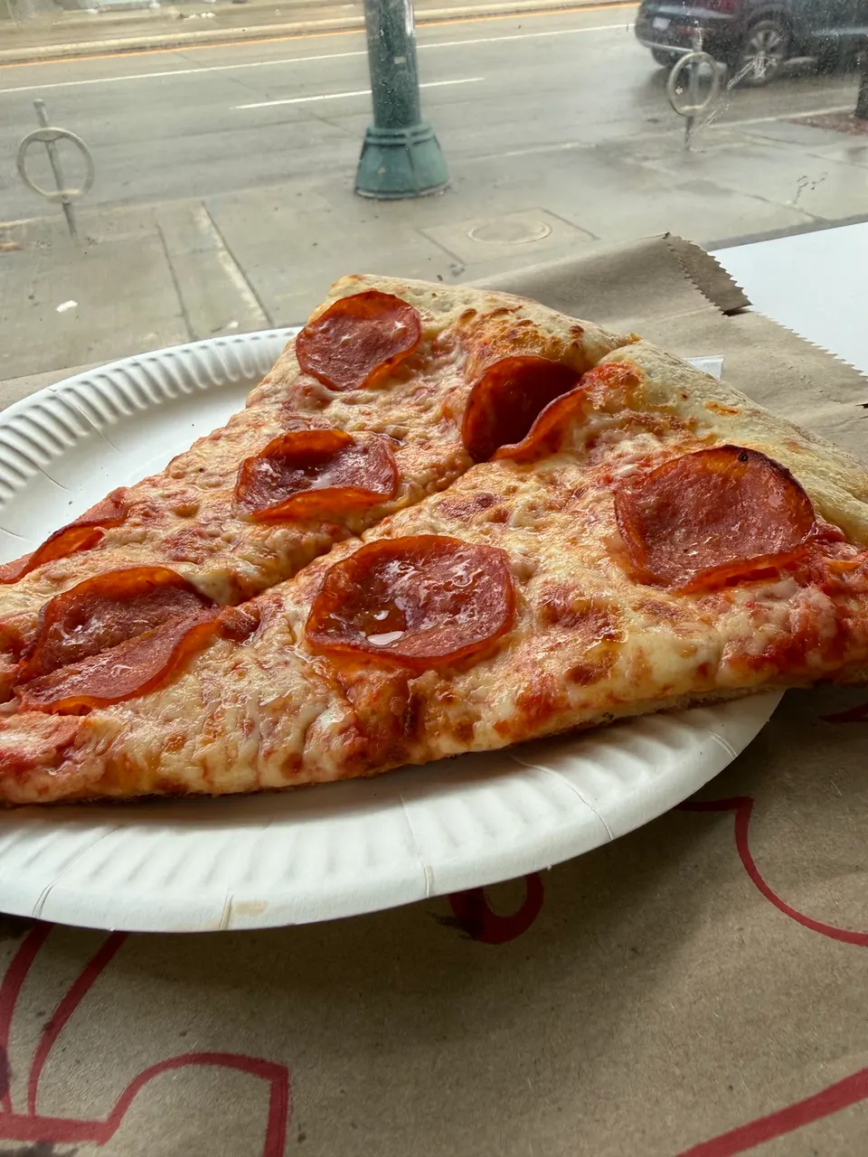 Two large slices of pepperoni pizza with melted cheese and crispy crust served on a white paper plate near a window.
