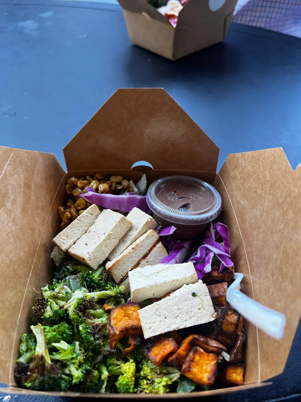 A brown cardboard takeout box filled with a healthy bowl containing sliced tofu, roasted sweet potatoes, broccoli florets, red cabbage, corn, and a small container of dressing.