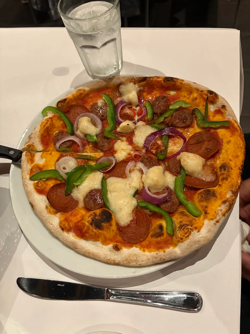 A freshly baked thin-crust pizza topped with pepperoni, sausage, green bell peppers, red onions, and melted mozzarella cheese, served on a white plate next to a glass of water and a knife.