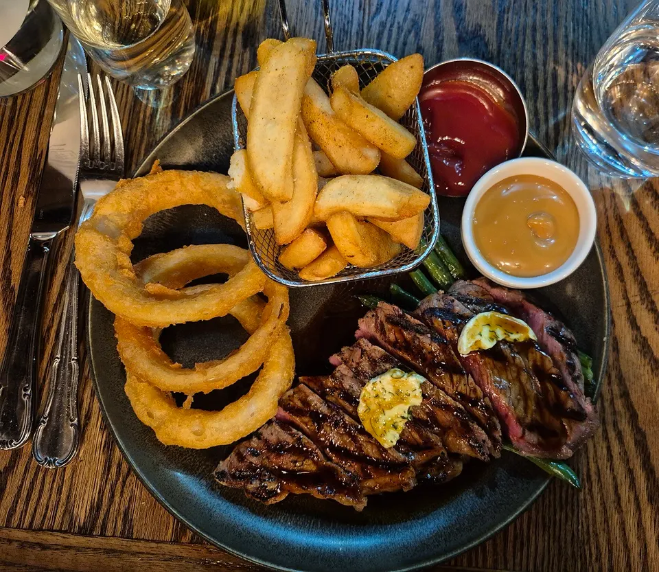 A top-down view of a steak dinner served on a dark plate, featuring sliced grilled steak topped with herb butter, thick-cut onion rings, a basket of potato wedges, ketchup, and a creamy dipping sauce.