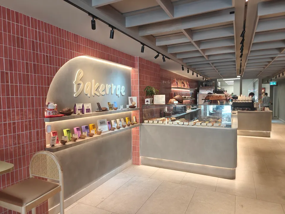 Modern bakery interior featuring a curved concrete display wall with 'Bakerrae' signage, pink tiled walls, a glass pastry counter, and warm under-counter lighting.