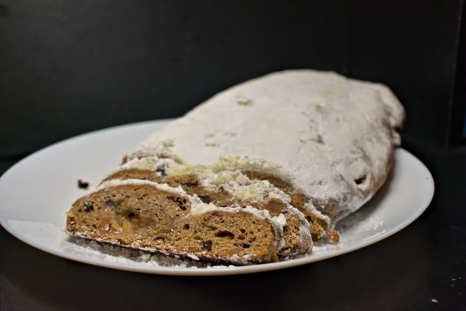A sliced loaf of German Stollen bread dusted with powdered sugar, served on a white plate against a dark background.