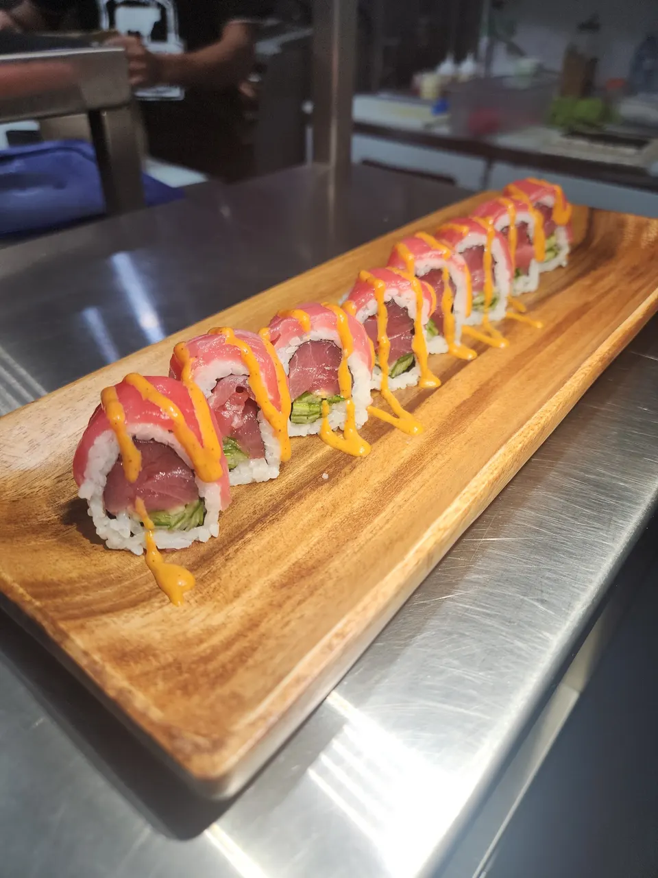 A row of eight sushi rolls topped with raw tuna and drizzled with spicy orange sauce, served on a long wooden platter on a stainless steel counter.