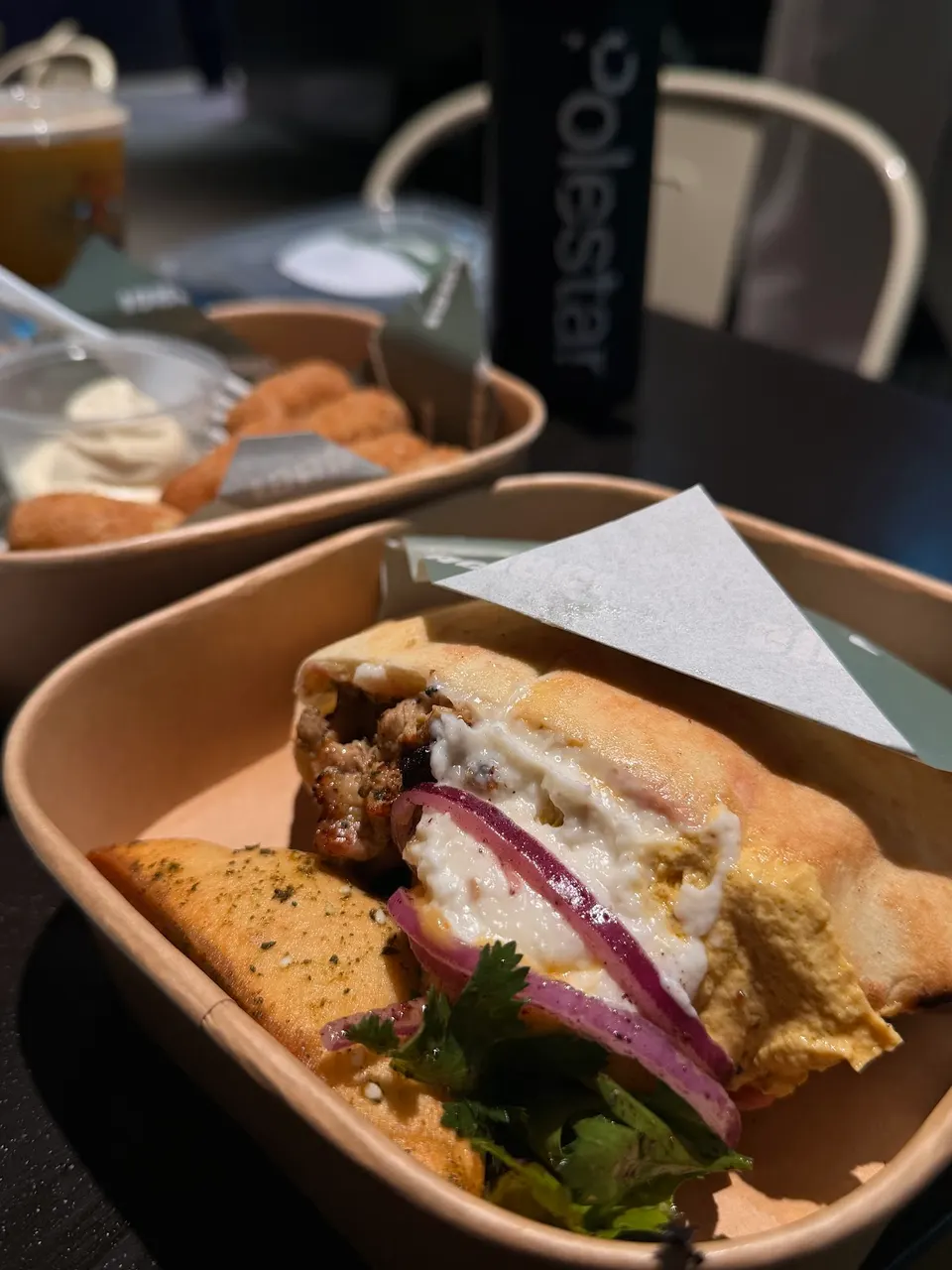 Close-up of a pita bread wrap filled with meat, white sauce, red onions, and cilantro, served in a light brown disposable container. Another similar container with fried food and a dip is blurred in the background, along with a black bottle with 'polestar' written on it.