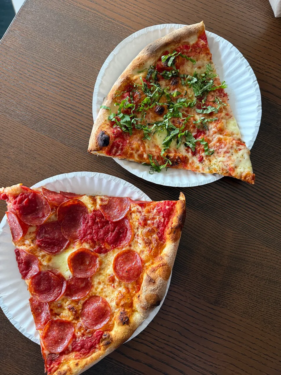 Two large slices of New York-style pizza served on white paper plates on a dark wooden table. One slice is topped with pepperoni and red sauce, and the other is topped with fresh shredded basil and cheese.