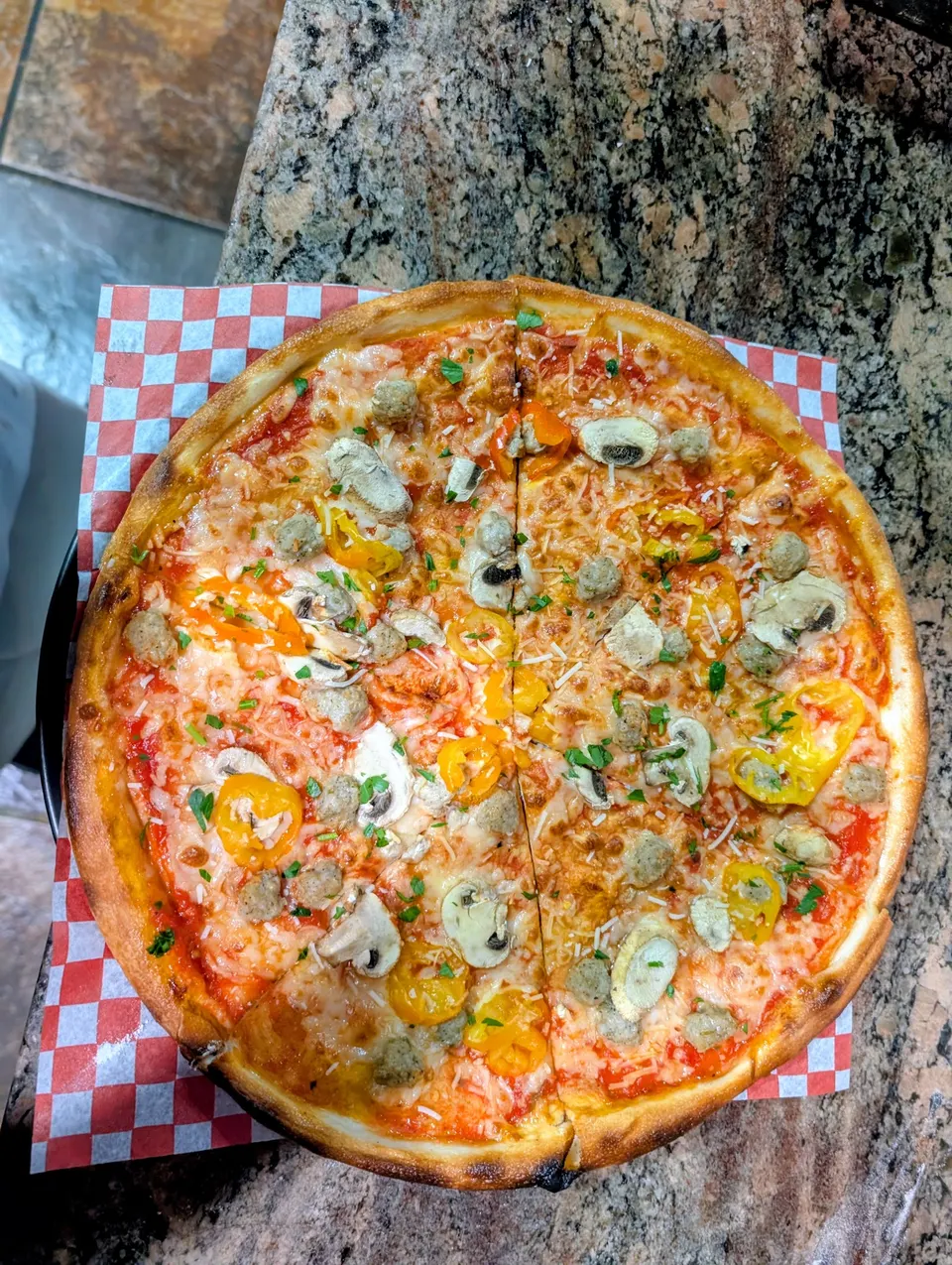 A freshly baked thin-crust pizza topped with sliced mushrooms, sausage chunks, yellow banana peppers, and fresh herbs, served on red and white checkered paper.