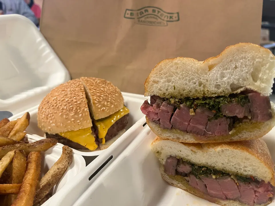 A sliced steak sandwich with green sauce, a sliced cheeseburger with melted cheese, and french fries are presented in white takeout containers, with a brown paper bag featuring a logo in the background.