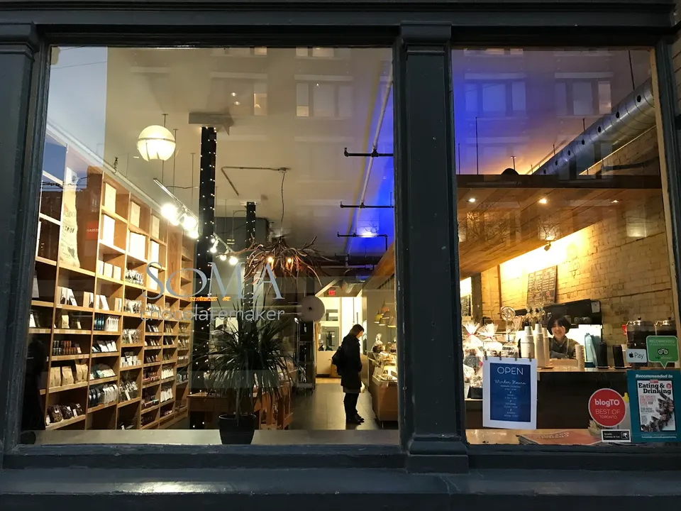 A view through a large glass window of SOMA Chocolatemaker, showing wooden shelves stocked with chocolate products, a potted plant, and a customer standing inside the warm, brightly lit shop.