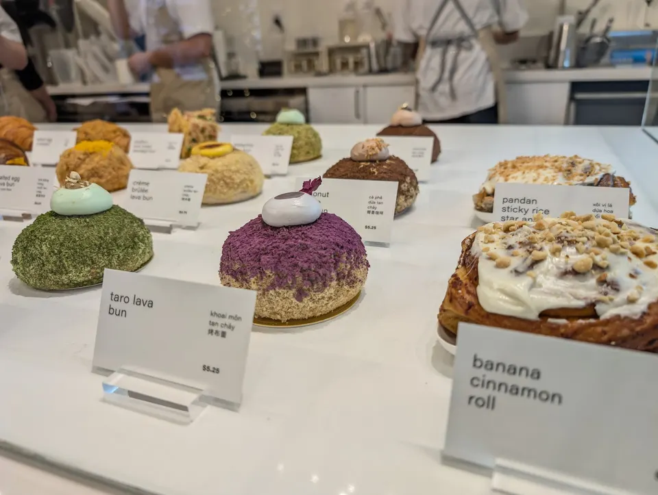 A display case at LA LA Bakeshop featuring various pastries like taro lava bun, banana cinnamon roll, and pandan sticky rice star anise, each with its own price tag.