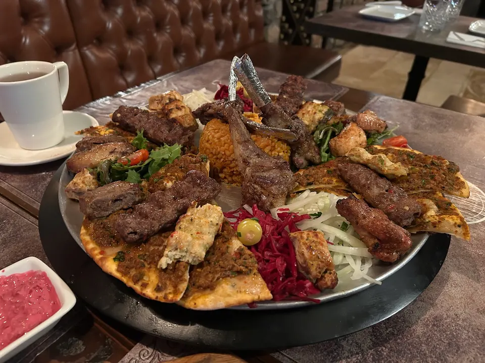 A large platter of mixed Turkish grilled meats including lamb chops, kebabs, and chicken skewers, served with bulgur rice, flatbread, red cabbage, and onions on a restaurant table.