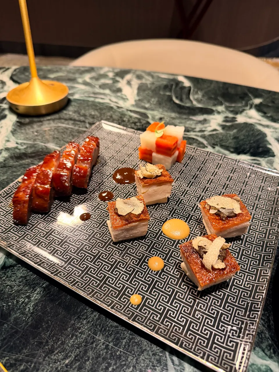 A square patterned plate featuring sliced glazed pork belly, crispy pork belly cubes topped with shaved truffle, and a small stack of pickled vegetables.