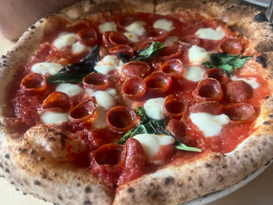 A freshly baked Neapolitan-style pizza with a charred, puffy crust, topped with bright red tomato sauce, melted fresh mozzarella pearls, crispy cupped pepperoni slices, and fresh basil leaves.