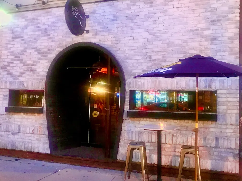 Exterior of Zitto Zitto Taverna featuring a light brick facade, a large arched doorway, a small outdoor table with two metal stools under a purple umbrella, and a circular sign with a hand gesture logo.