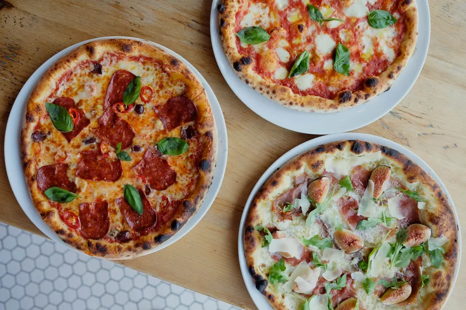 Three different wood-fired pizzas served on white plates, arranged on a light wooden table. One pizza has pepperoni and basil, one is a classic Margherita, and the third features prosciutto, figs, and arugula.