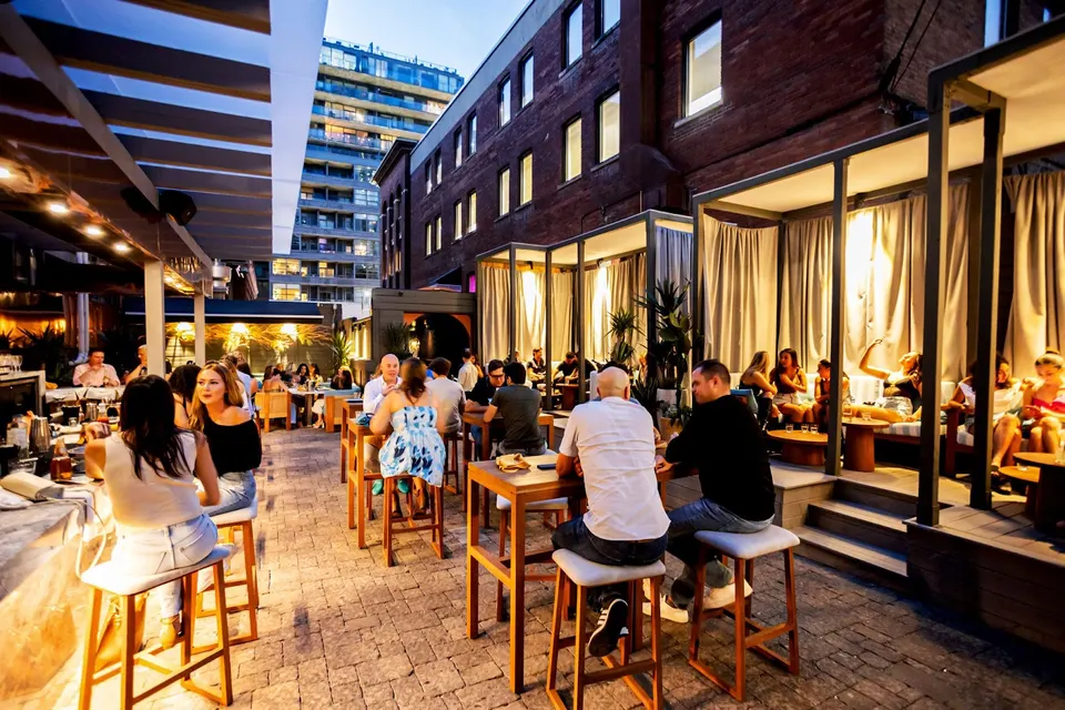 A bustling outdoor patio at night with patrons seated at high-top tables and in private curtained cabanas, set against a backdrop of brick buildings and city lights.