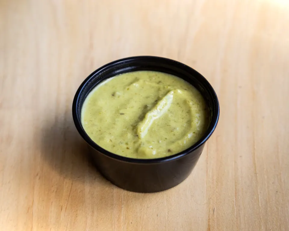 A small black plastic cup filled with a creamy green sauce, likely a herb-infused dipping sauce, sitting on a light wooden table.
