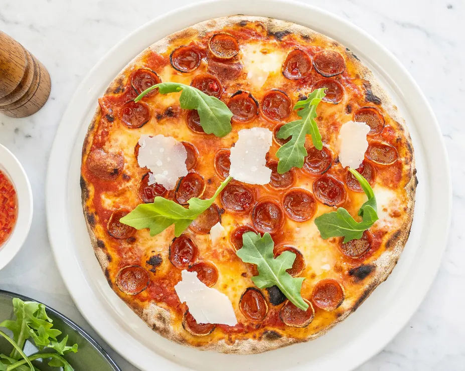 A freshly baked thin-crust pepperoni pizza topped with shaved parmesan cheese and fresh arugula leaves, served on a white plate.