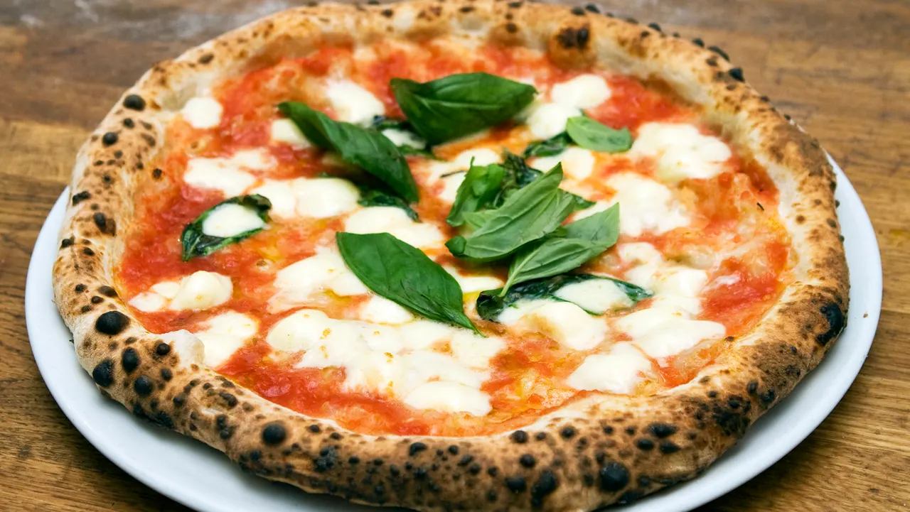 A freshly baked Neapolitan-style Margherita pizza on a white plate, featuring a charred, puffy crust, melted mozzarella cheese, tomato sauce, and fresh green basil leaves.