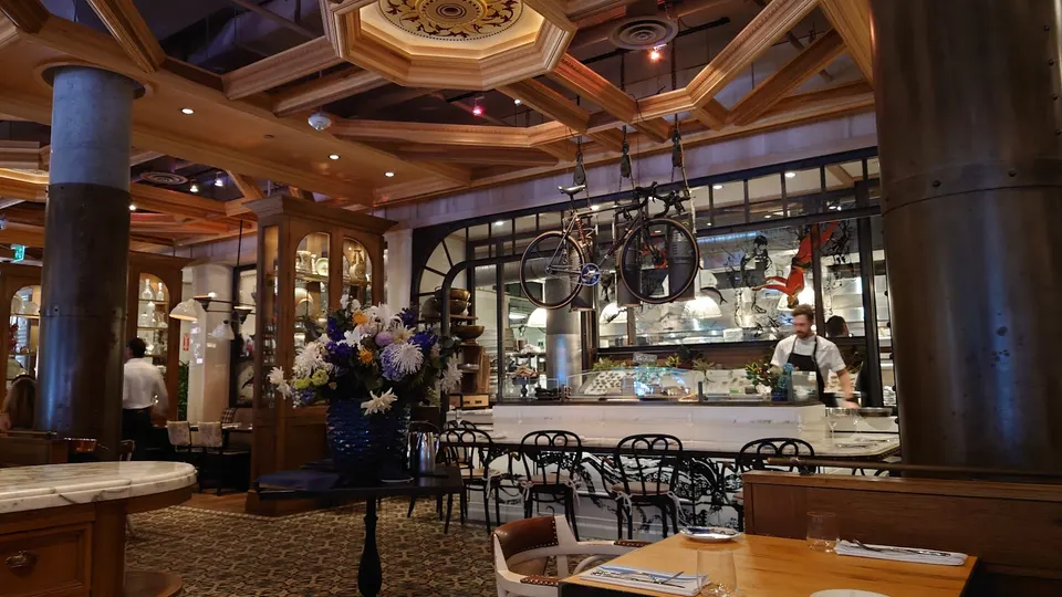 Interior view of Cluny Bistro & Boulangerie showing a dining area with wooden tables, a large floral arrangement, a bar counter with stools, and a bicycle suspended from the ceiling.