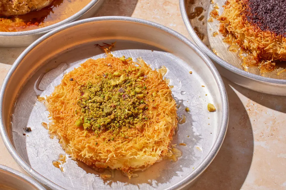 A round, golden-brown knafeh pastry topped with crushed green pistachios, served in a round metal baking pan with syrup.