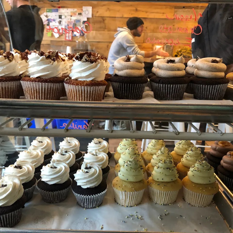 A bakery display case filled with rows of freshly baked cupcakes, including carrot cupcakes with white frosting and nuts, chocolate cupcakes with mocha frosting, and vanilla cupcakes with yellow frosting and sprinkles.