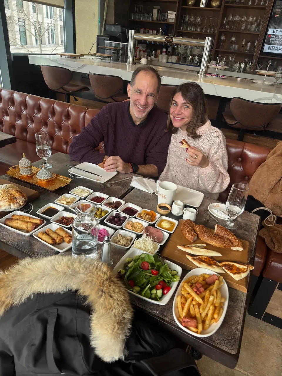 A smiling couple sits at a restaurant table filled with a Turkish breakfast spread, including simit, various small dipping dishes, salad, fries, and pastries.