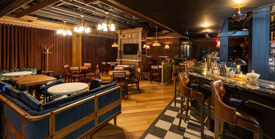 A dimly lit, stylish bar and lounge area with a long marble-topped bar, leather barstools, wooden dining tables, plush blue velvet booths, and warm vintage lighting.