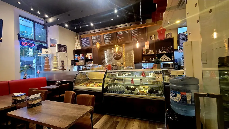 Interior of Melt n dip Sweets, Desserts and Gelato showing wooden tables and chairs, a red booth, and glass display cases filled with desserts and gelato under warm lighting.