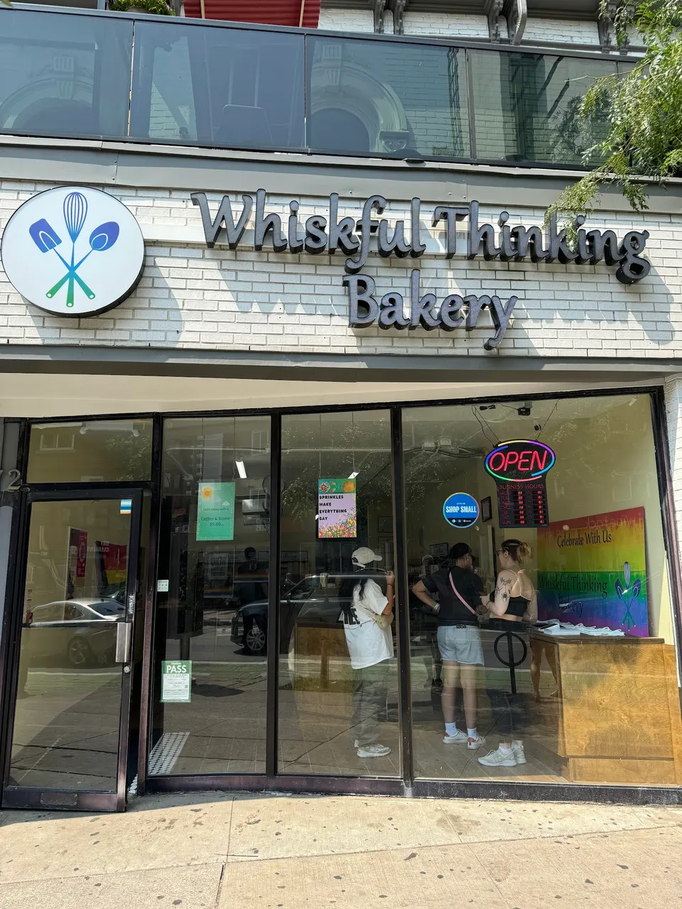 The storefront of Whiskful Thinking Bakery, featuring a white brick facade, a large sign with the bakery's name, a circular logo with baking utensils, and customers visible through the glass windows.