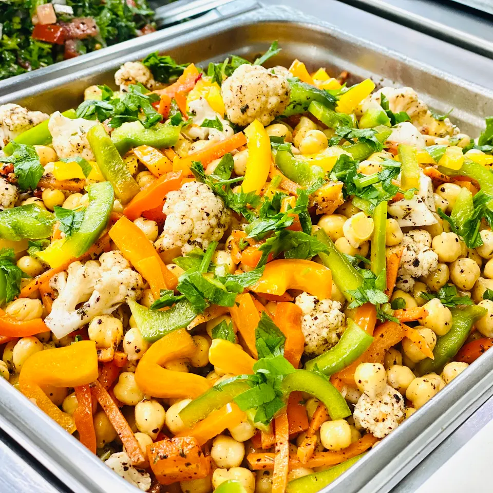 A close-up shot of a colorful chickpea salad with cauliflower florets, chopped yellow and orange bell peppers, green peppers, and fresh parsley in a metal serving tray.