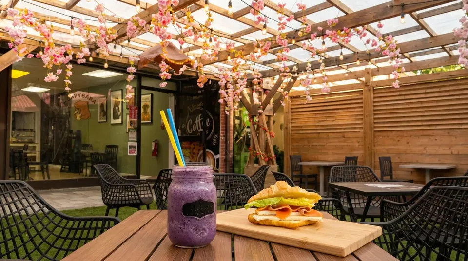 A purple smoothie in a mason jar with yellow and blue straws next to a croissant sandwich on a wooden board, set on an outdoor patio table under a wooden pergola decorated with pink cherry blossoms.