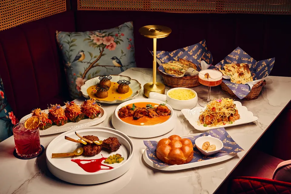 A marble restaurant table filled with a spread of Indian dishes, including curries, naan bread, appetizers, and colorful cocktails, set against a plush booth with floral pillows.