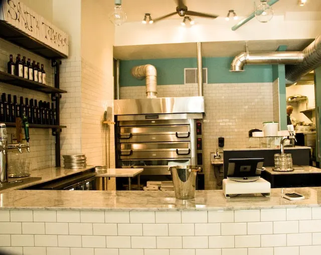 The interior of Salt & Tobacco Pizzeria showing a marble service counter, a large stainless steel pizza oven in the background, white subway tile walls, and shelves stocked with bottles.