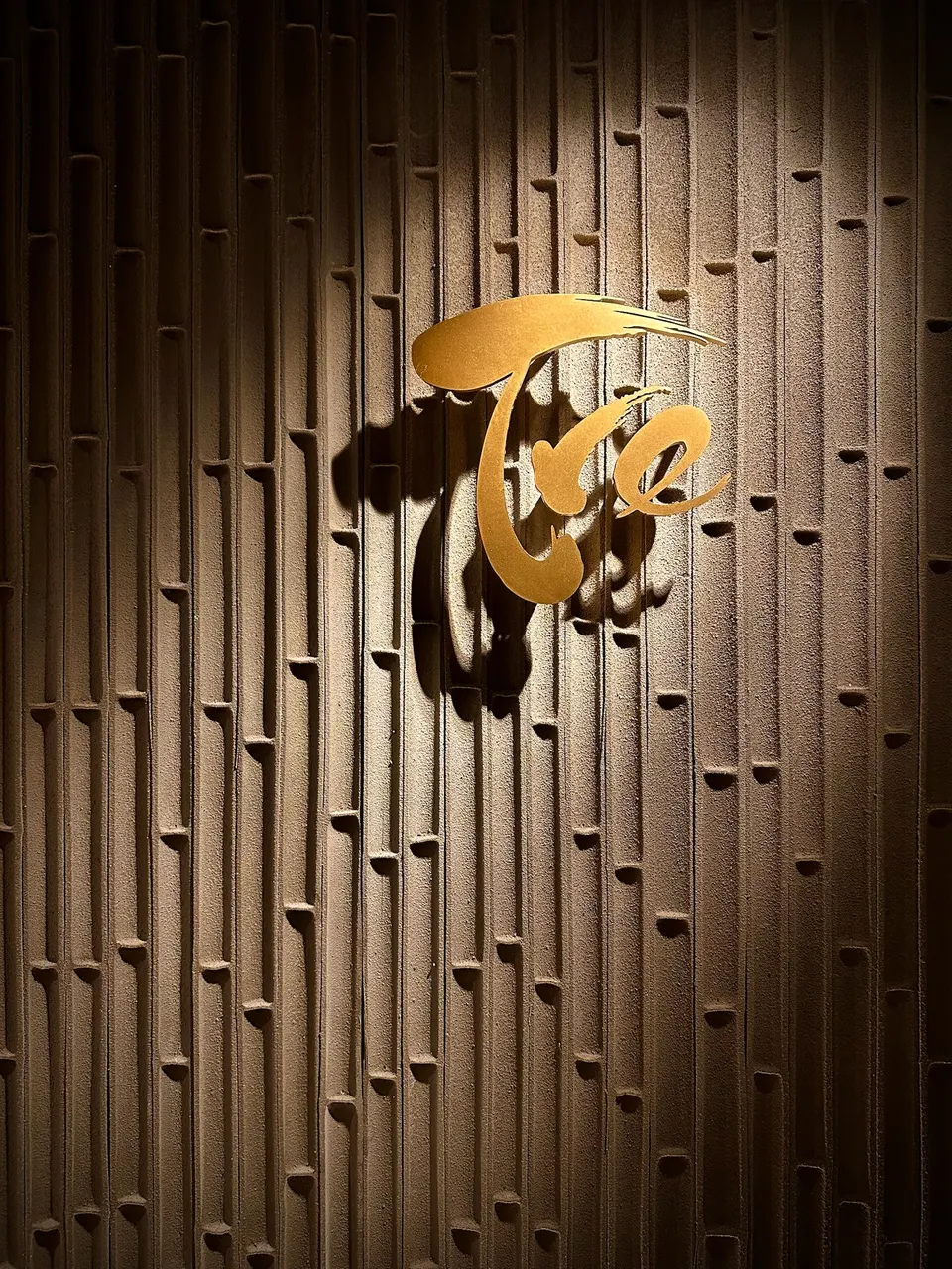 A golden metallic sign spelling 'Tre' in a stylized, flowing cursive font, mounted on a textured brown wall designed to look like vertical bamboo stalks under a spotlight.