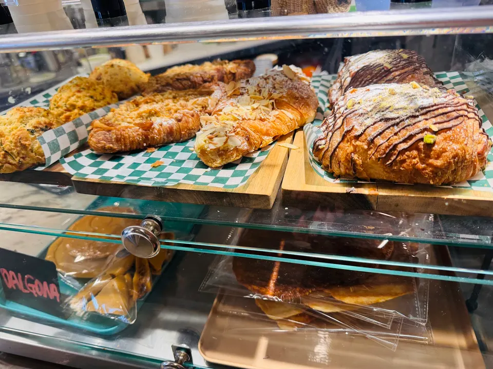 A glass display case filled with freshly baked pastries including almond croissants, chocolate-drizzled pastries, and scones on wooden trays lined with green checkered paper.