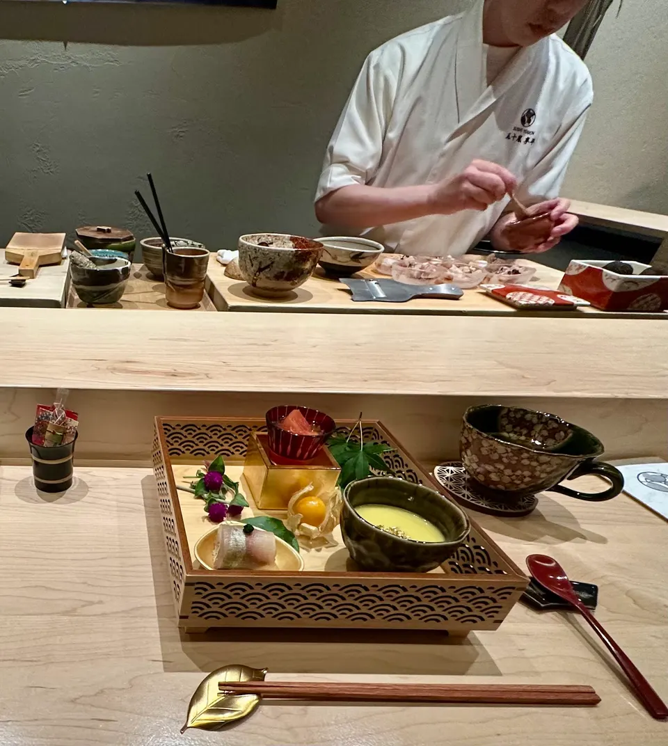 A chef prepares food behind a wooden counter while a beautifully arranged multi-course appetizer tray sits on the dining table in the foreground.
