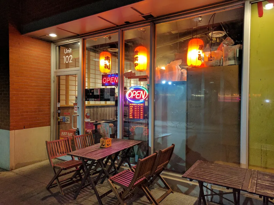 Exterior view of Kibo Sushi House at night, featuring a glass storefront with glowing red and orange paper lanterns, neon 'OPEN' signs, and wooden patio tables and chairs set up on the sidewalk.
