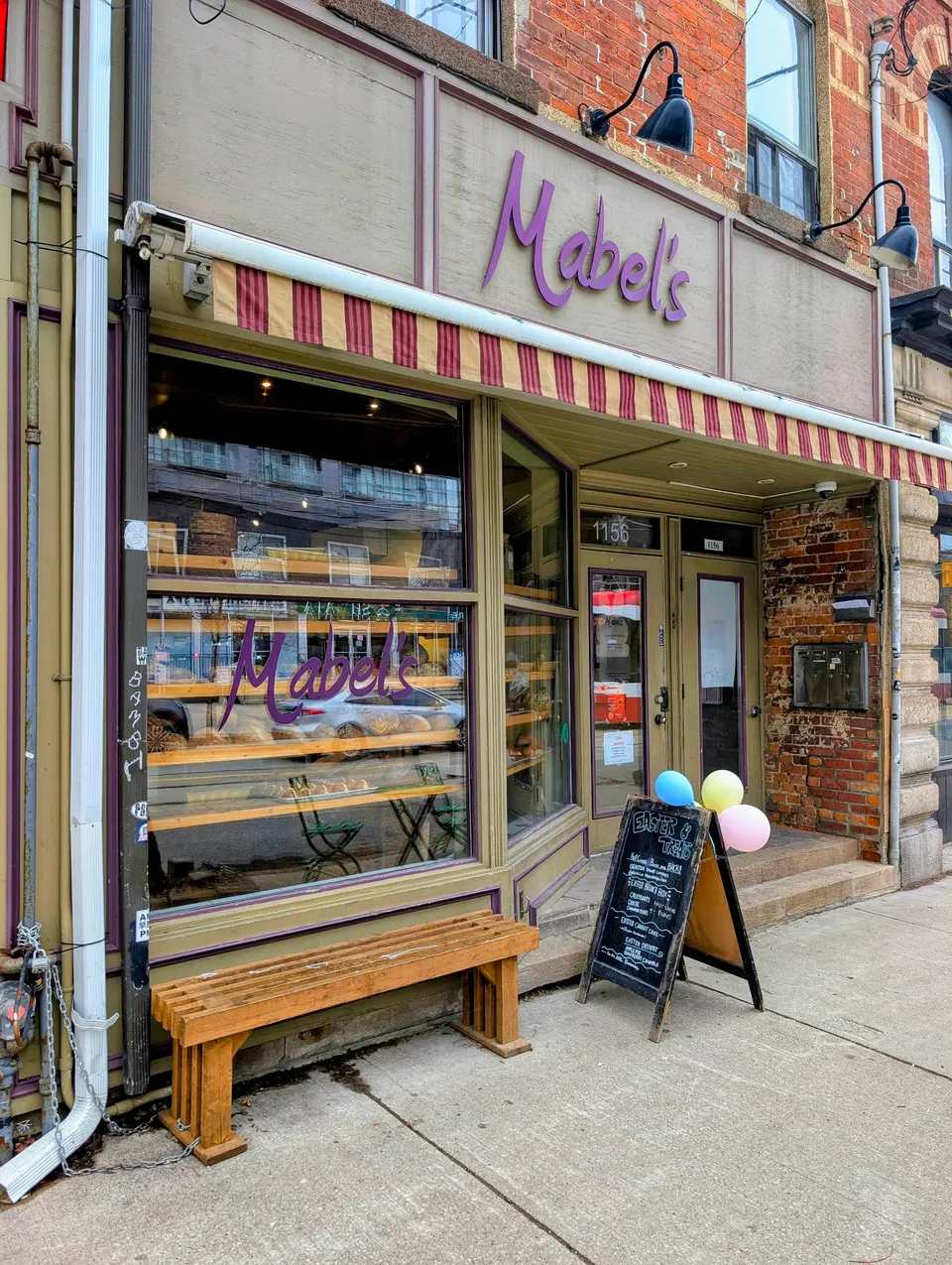 Exterior storefront of Mabel's Bakery with a purple sign, large display windows showing baked goods, a wooden bench outside, and a chalkboard menu sign decorated with balloons on the sidewalk.