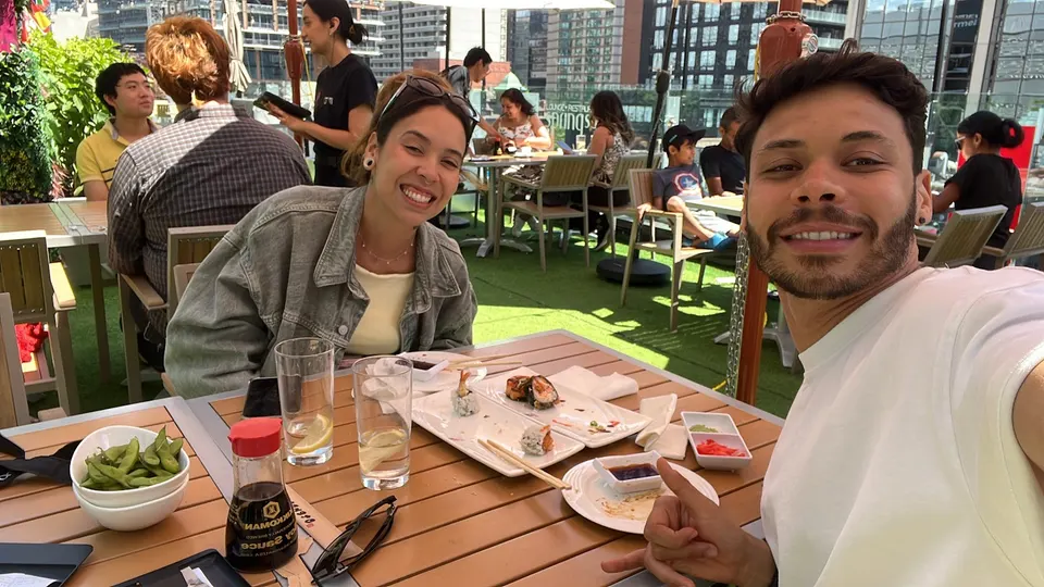 A smiling young couple takes a selfie at an outdoor rooftop patio table with sushi plates, edamame, and soy sauce, with other diners in the background.