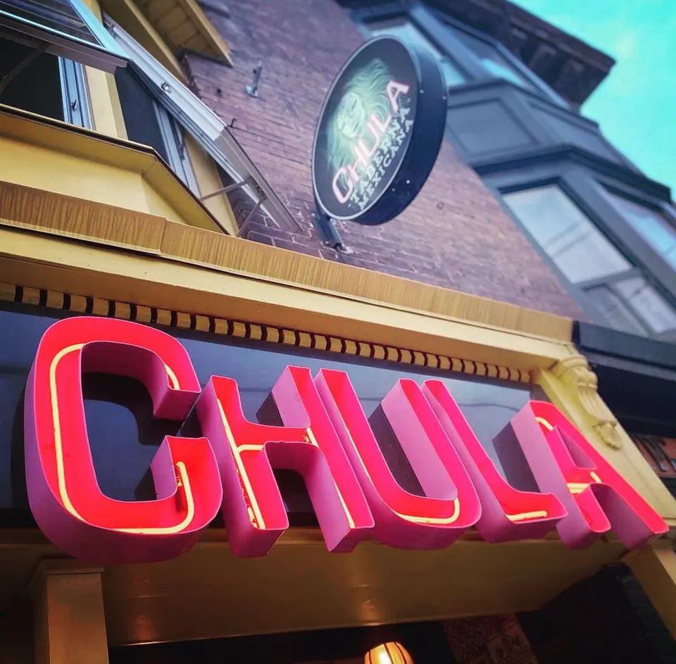 A close-up, low-angle shot of the vibrant pink and yellow neon sign spelling 'CHULA' on a storefront, with a circular hanging sign above reading 'CHULA TABERNA MEXICANA'.
