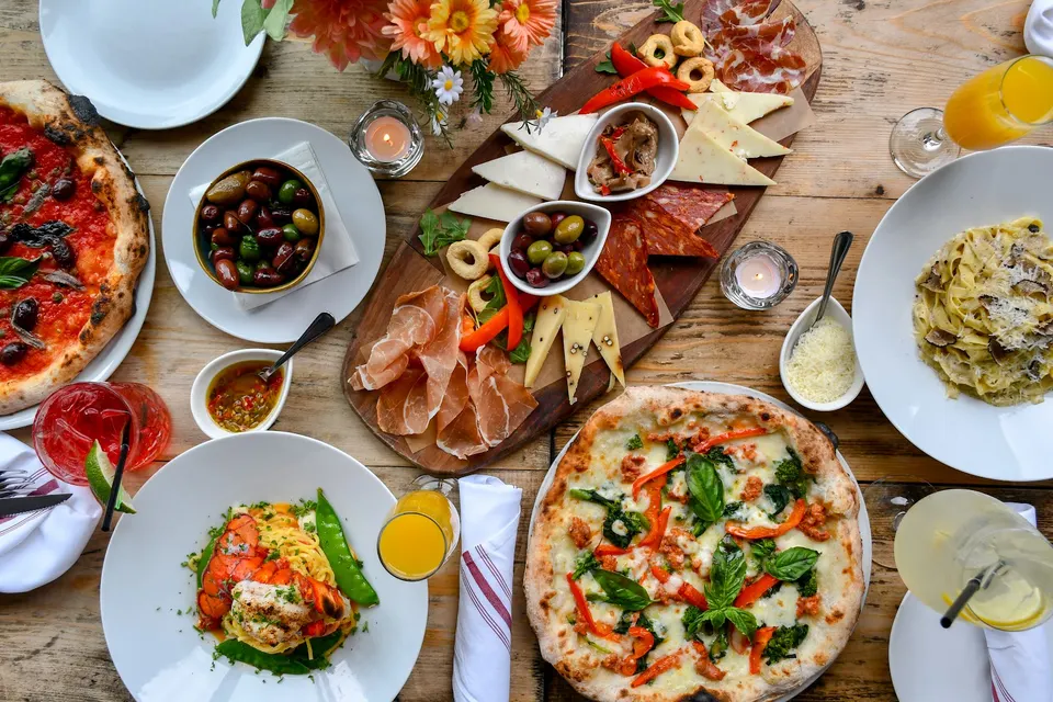 An overhead view of a rustic wooden table filled with Italian dishes including two pizzas, a pasta dish, a seafood pasta plate, a charcuterie board, olives, and cocktails.