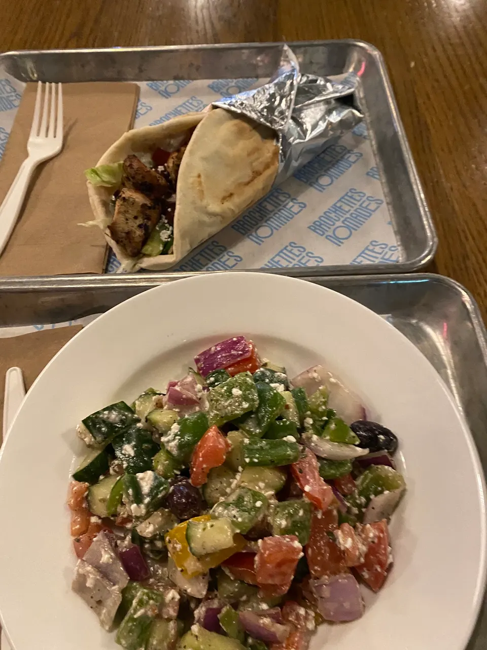 A Greek salad with cucumbers, tomatoes, red onions, olives, and feta cheese in a white bowl, next to a chicken gyro wrapped in pita and foil on a metal tray.