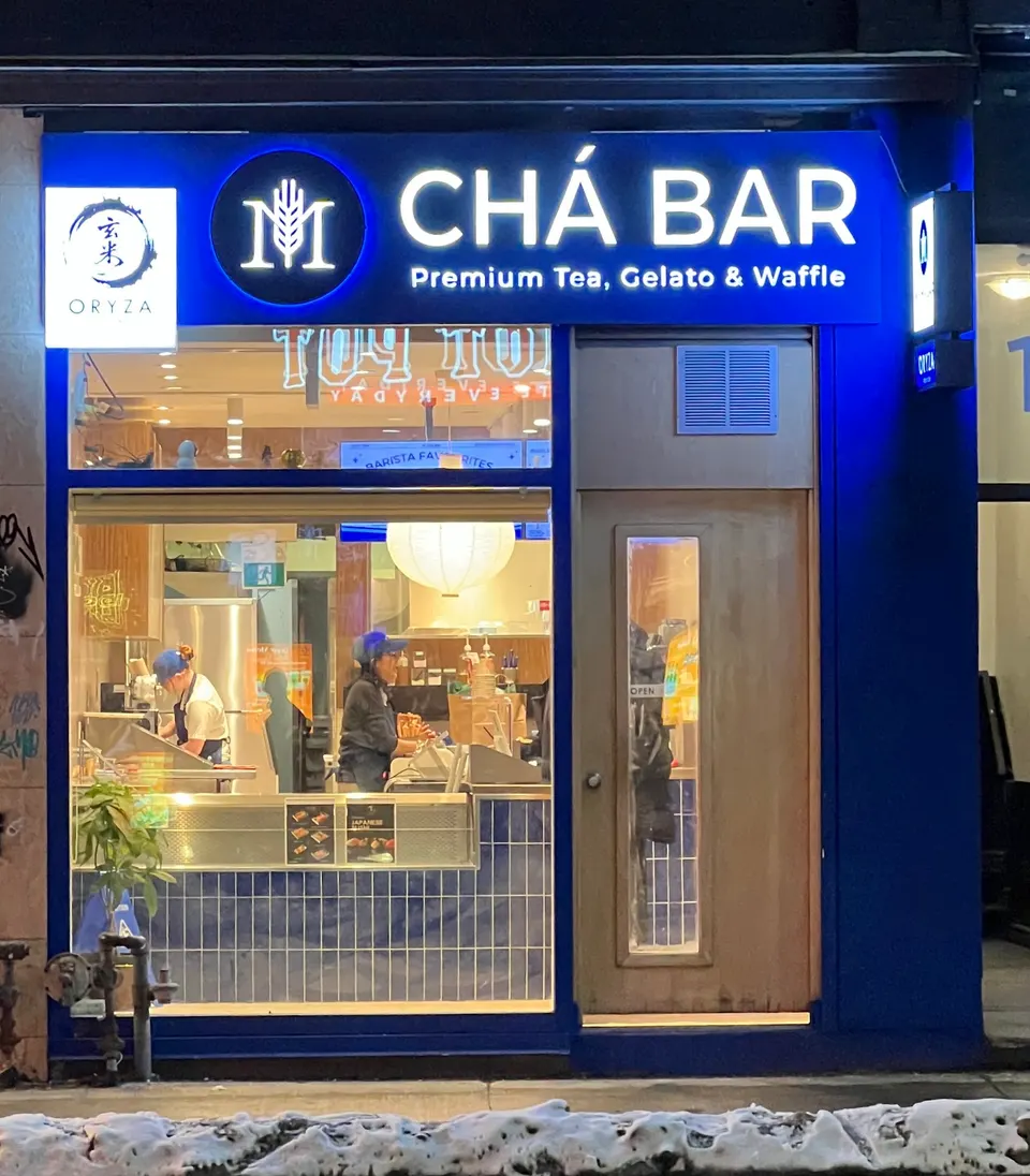 Exterior storefront of ORYZA Sushi Bar and Chá Bar at night, featuring a bright blue facade, illuminated signs, and a large window showing staff working inside.